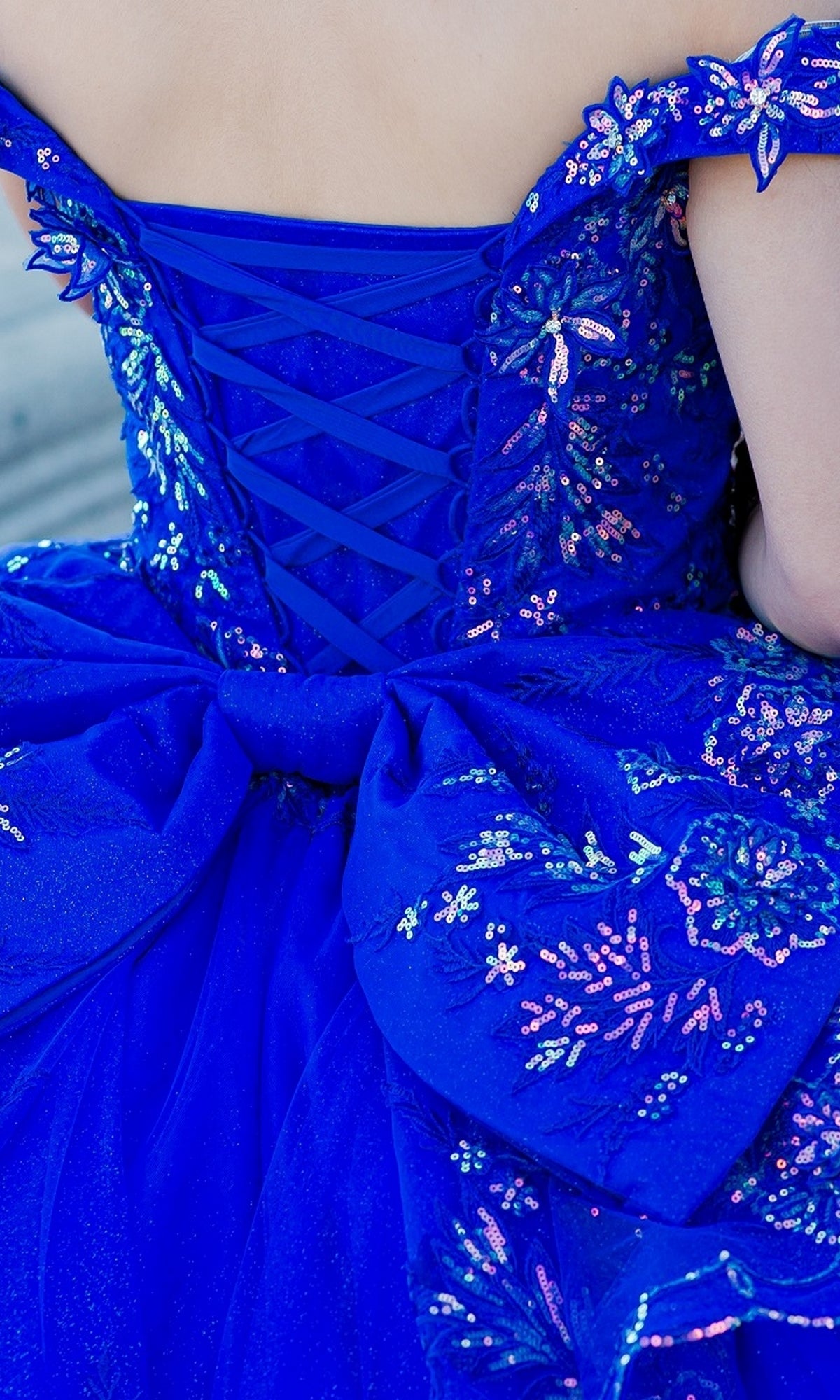 A person wearing the Quinceanera Ball Gown 8100J: a bright blue, off-the-shoulder dress featuring sequin floral patterns, iridescent beaded details, a lace-up back, and a large decorative bow.